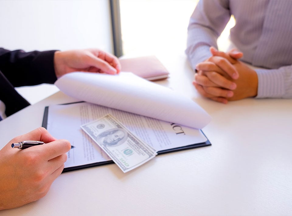 Dos personas en un escritorio durante una transacción financiera relacionada con el consumo, uno con documentos y el otro con las manos entrelazadas, con dinero en efectivo y un bolígrafo visibles sobre la mesa.