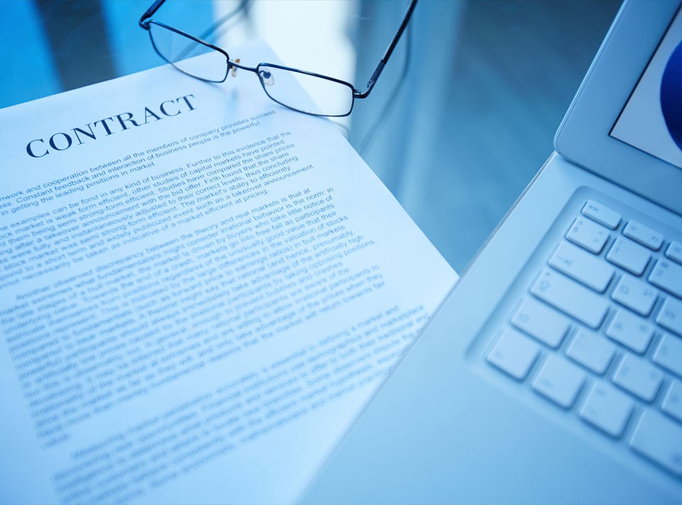 Un primer plano de un contrato mercantil impreso sobre un escritorio con anteojos encima, junto a una computadora portátil abierta con una luz en tonos azules reflejada, creando un ambiente profesional.