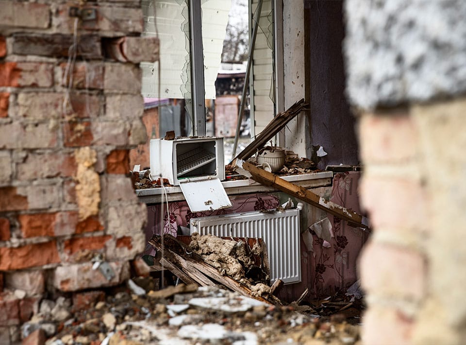 Vista a través de una pared de ladrillos rota que muestra el interior de una vivienda dañada, con un radiador derribado y escombros.