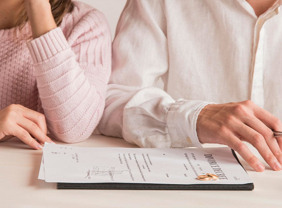 Dos personas sentadas en una mesa, una con un suéter rosa y la otra con una camisa blanca, con las manos apoyadas cerca de un documento titulado "acuerdo de divorcio", discuten sus opciones con un abogado de la familia.