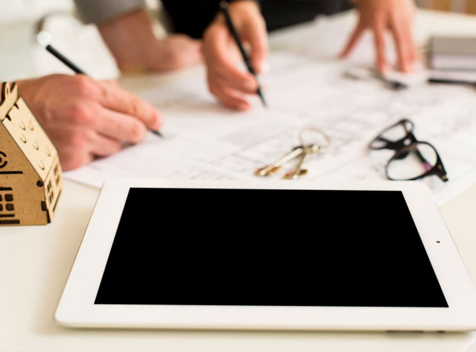 Primer plano de un espacio de trabajo concursal con manos escribiendo en planos arquitectónicos, una tableta digital con pantalla negra, modelo de casa, llaves y vasos sobre la mesa.