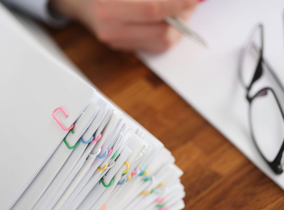 Primer plano de separadores de pestañas de colores en una carpeta con una persona escribiendo sobre consumo económico al fondo y un par de vasos apoyados sobre la mesa.