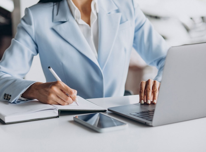 Una mujer profesional con una chaqueta azul toma notas en una libreta mientras trabaja en una computadora portátil, con un teléfono inteligente colocado a su lado en el escritorio. La descripción proporcionada es insuficiente para identificar palabras clave SEO relevantes, ya que es demasiado breve y carece de contexto o información relacionada con el contenido.