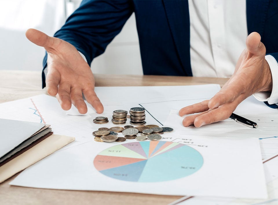 Las manos de un hombre de negocios gesticulando sobre gráficos financieros con pilas de monedas dispuestas sobre ellos, indicando diferentes segmentos de un gráfico circular sobre una mesa para analizar las tendencias de consumo.